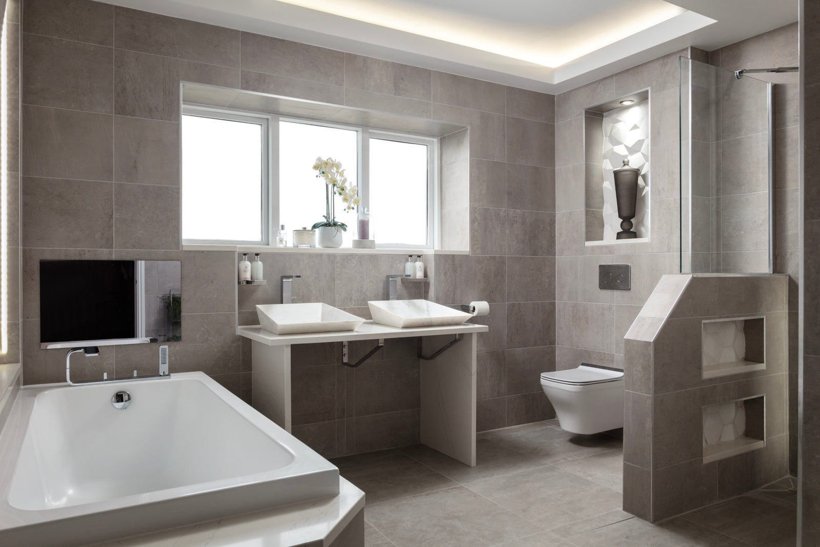 Lisa Hensby's own master bathroom with warm grey large-format tiles, a built-in bath, twin basins under the window and a walk-in shower behind a half-height wall