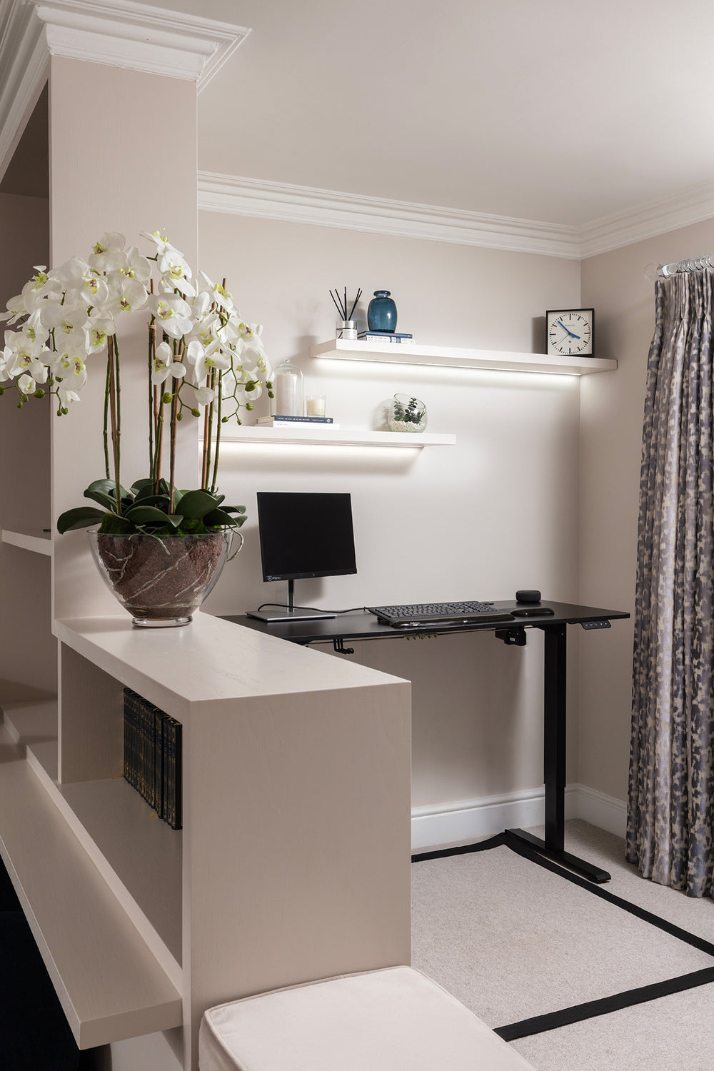 Compact home office behind a half-height partition, with a black desk and lit floating shelves, by Lisa Hensby Design & Build Studio.