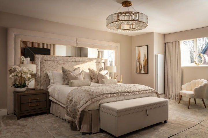 A luxurious neutral bedroom with a tall upholstered headboard, large mirror, layered champagne bedding, a gold crystal chandelier, and a cream bouclé armchair in the corner - designed by Lisa Hensby Design & Build Studio.