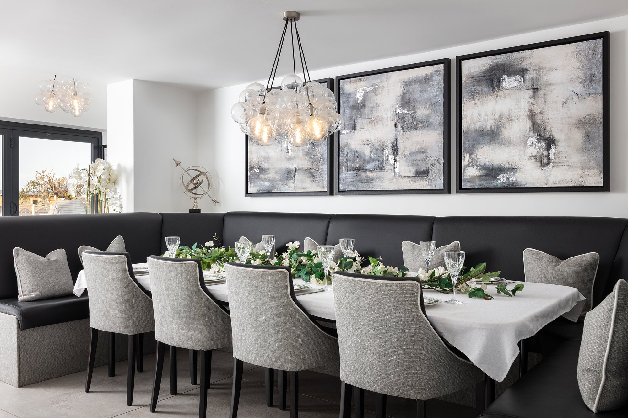 An elegant monochrome dining room featuring a curved black leather banquette, grey upholstered dining chairs, a statement bubble glass chandelier and a pair of large abstract artworks in muted greys and cream — designed by Lisa Hensby Design & Build Studio.