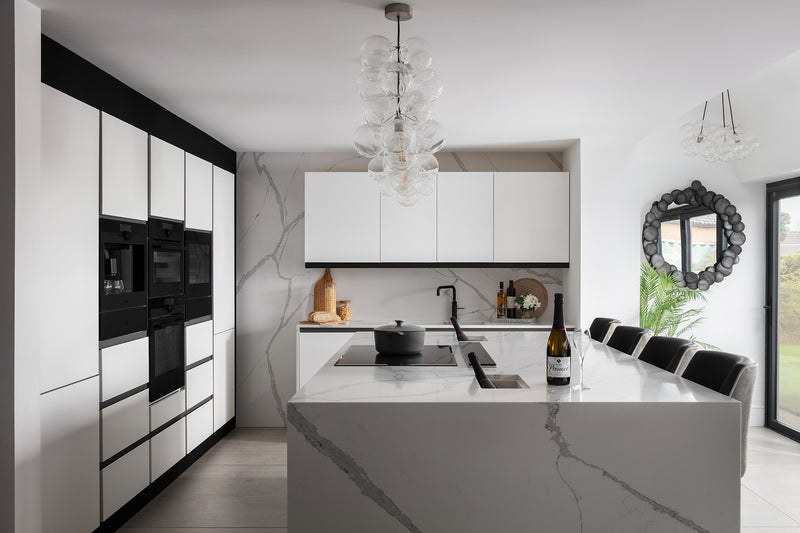 Monochrome handleless kitchen with full-height white cabinetry, marble-effect quartz island and splashback, black integrated appliances and bubble glass pendant light. Designed by  Lisa Hensby Design & Build Studio.