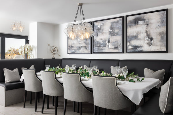 Long dining booth with a charcoal banquette, grey chairs and a glass bubble pendant, set beneath three abstract canvases with the kitchen beyond. Designed by Lisa Hensby  Design & Build Studio.