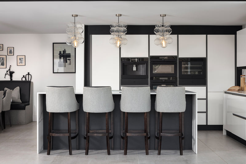 Kitchen island with four grey bar stools and three glass bubble pendants, set against white and black cabinetry with integrated appliances. Designed by Lisa Hensby Design & Build Studio.