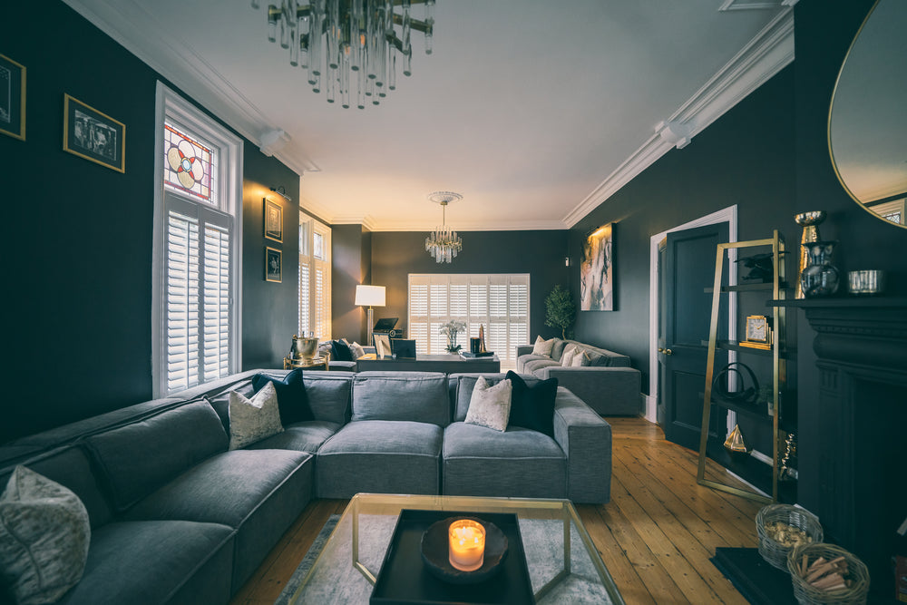 Grey modular sofa around a glass and brass coffee table in a dark-walled period living room with white plantation shutters, by Lisa Hensby Design & Build Studio.