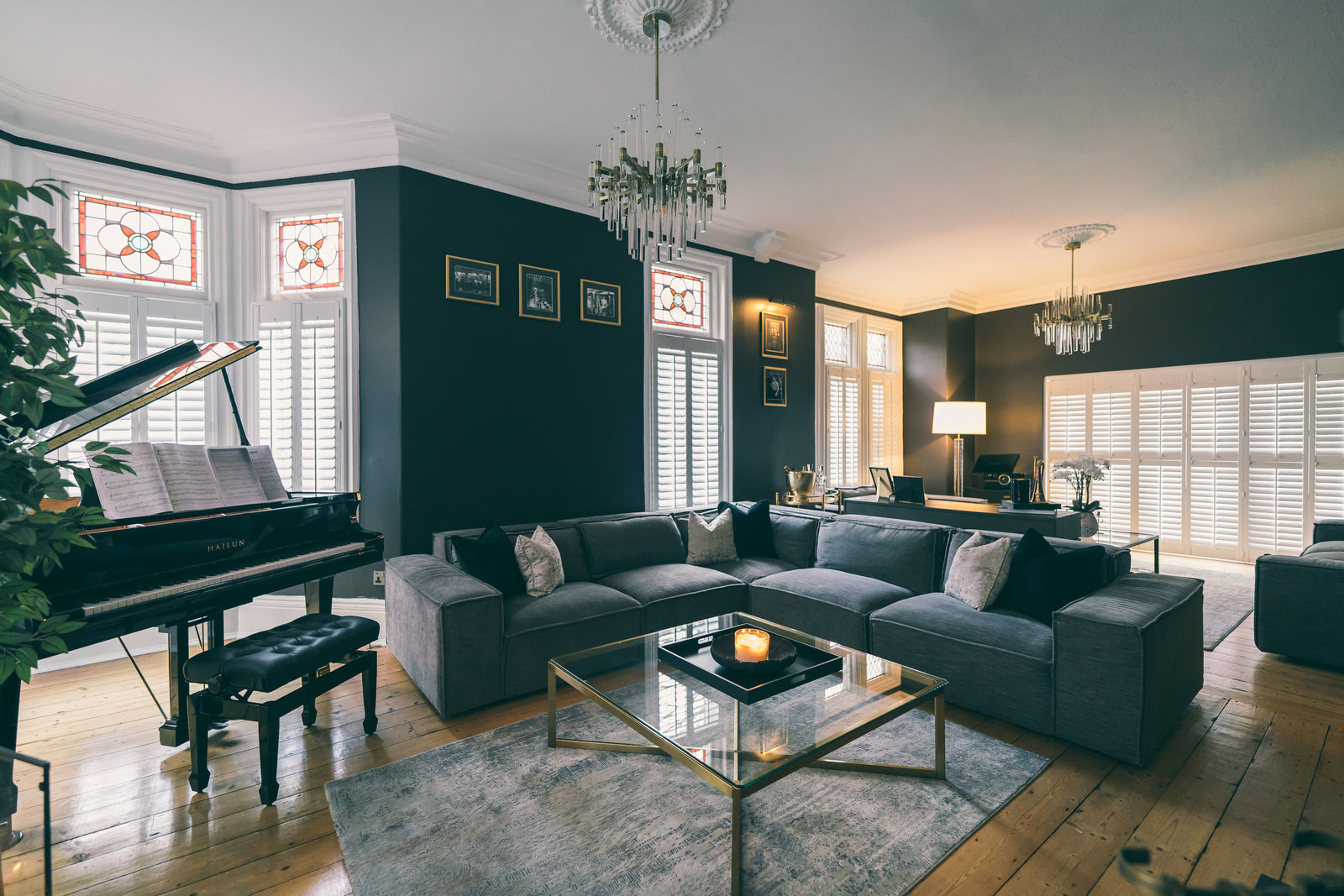 Period living room in Manchester by Lisa Hensby Design & Build Studio, with near-black walls, stained-glass bay windows, a grand piano, glass-drop chandeliers and a grey modular sofa.