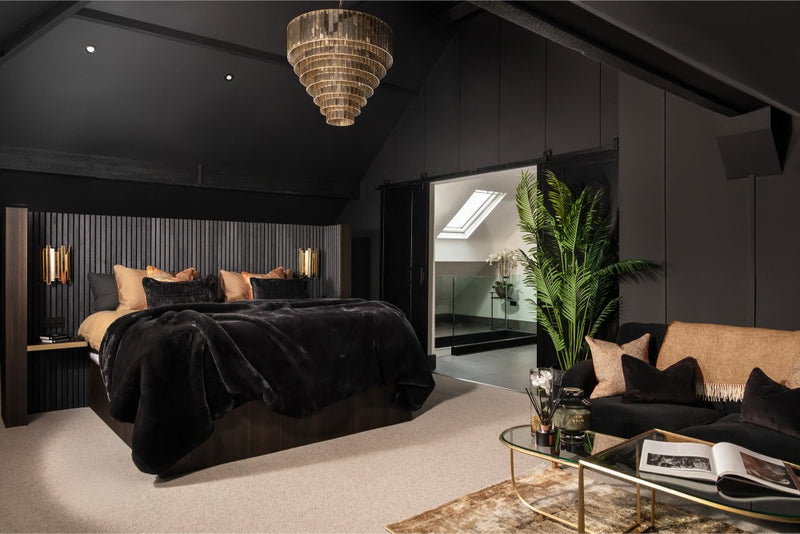 A dramatic dark loft bedroom with a black faux fur bedspread, vertical slatted headboard wall, tiered gold chandelier, and an open ensuite bathroom beyond a sliding barn door - designed by Lisa Hensby Design & Build Studio.