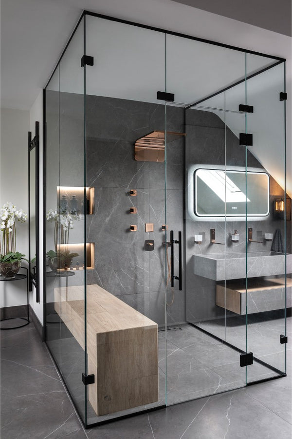 Frameless glass-cube shower lined in grey porcelain with copper fittings, a wood-effect bench and illuminated niches, beside a stepped vanity and backlit mirror.