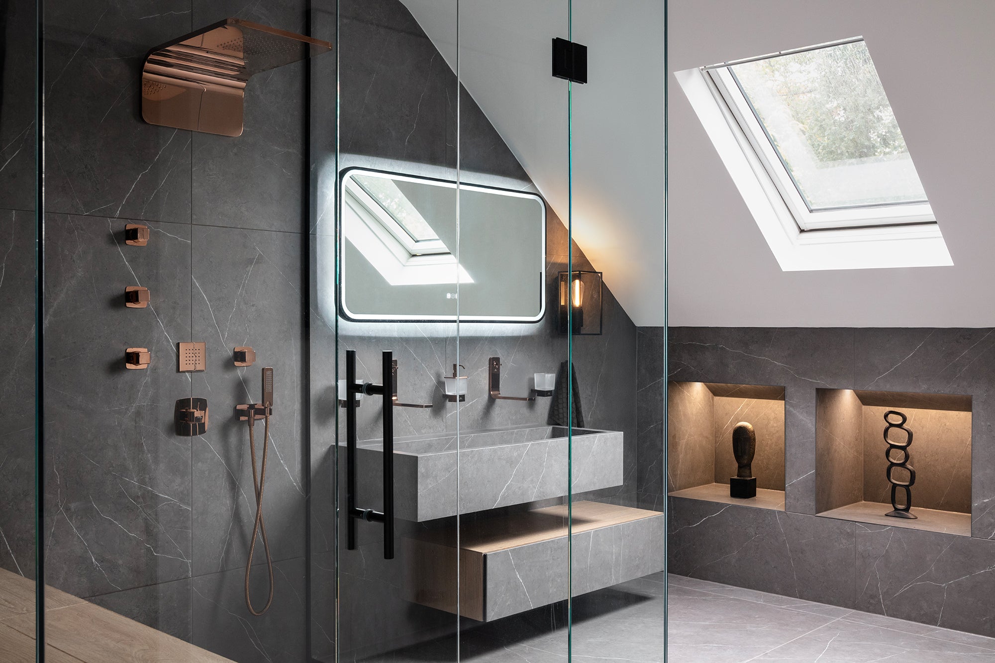 A luxury loft ensuite bathroom with dark grey marble-effect tiles, rose gold shower fittings, a wall-mounted stone basin, LED backlit mirror and Velux skylight - designed by Lisa Hensby Design & Build Studio.