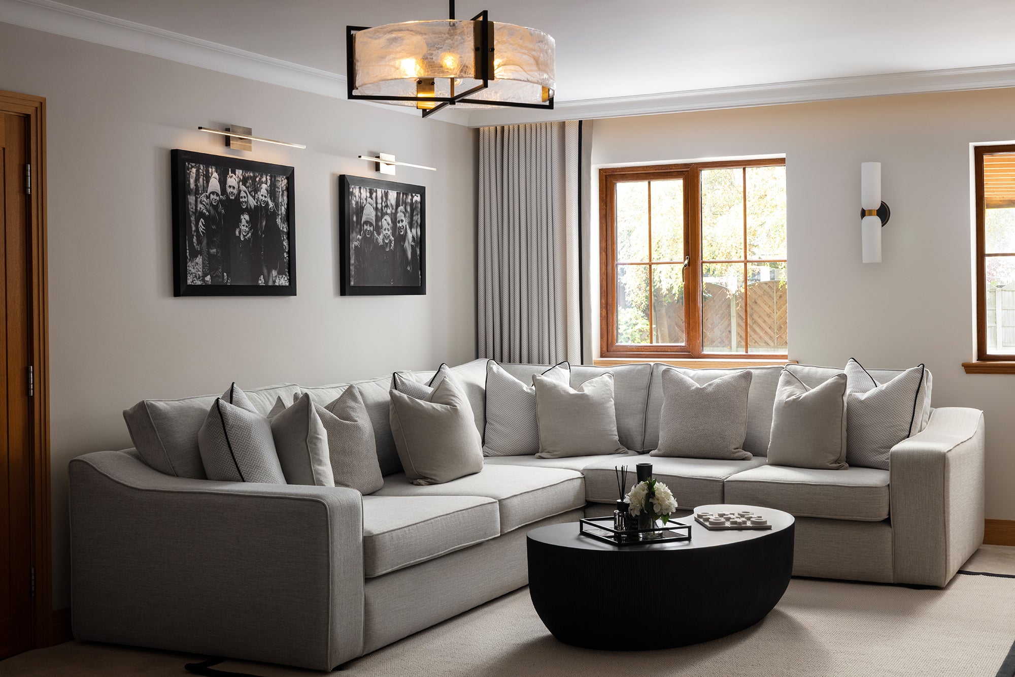 A neutral living room featuring a large grey corner sofa with black piping, round black oval coffee table, frosted glass pendant light, wall sconces and black and white framed photography - designed by Lisa Hensby Design & Build Studio.