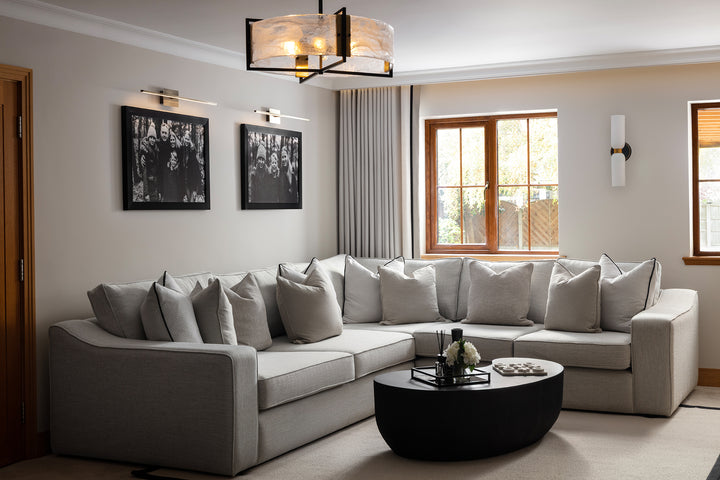 A neutral living room featuring a large grey corner sofa with black piping, round black oval coffee table, frosted glass pendant light, wall sconces and black and white framed photography - designed by Lisa Hensby Design & Build Studio.