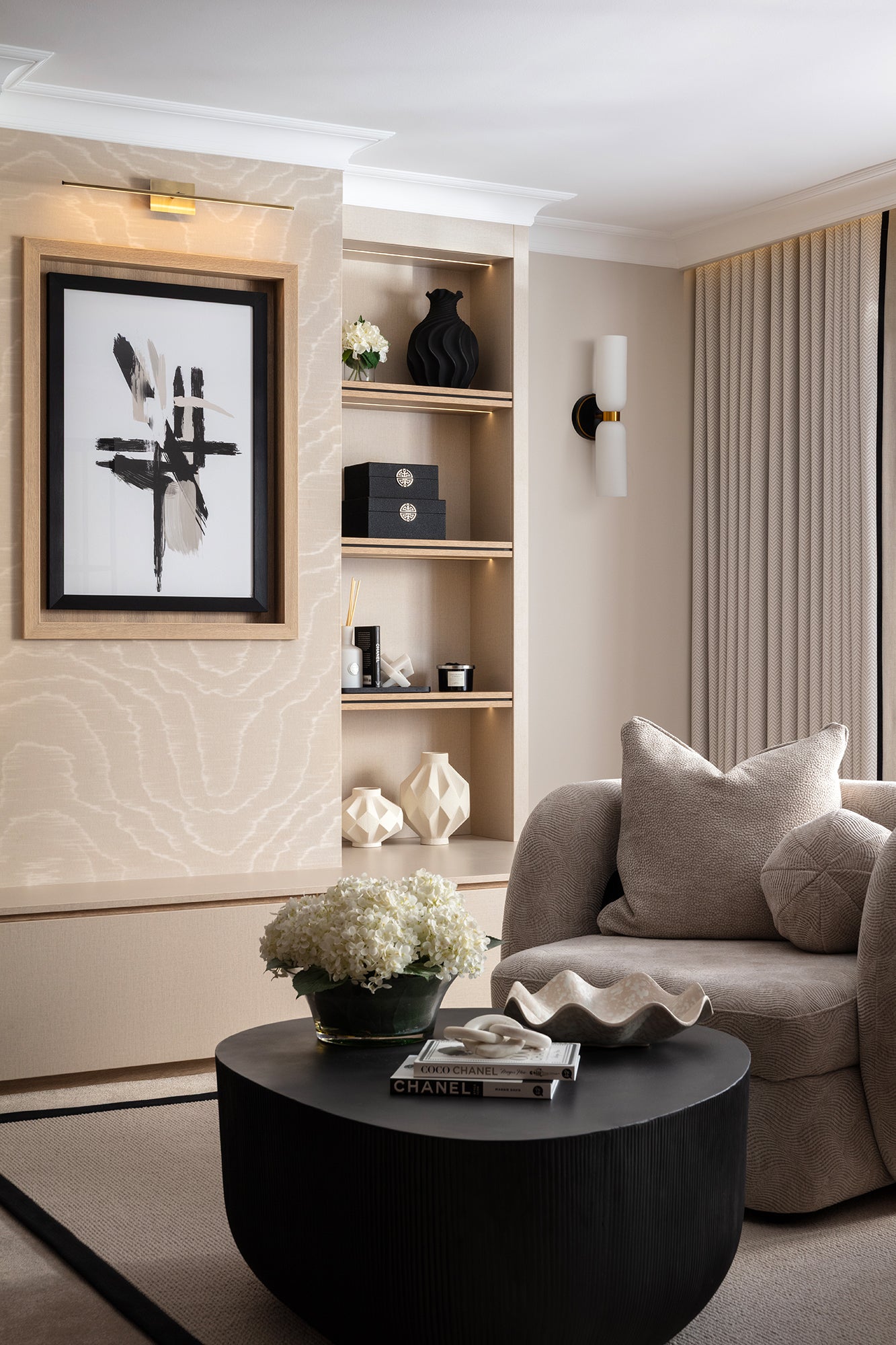 A refined sitting room corner with a dark ribbed drum coffee table, a taupe bouclé armchair, built-in shelving styled with black and cream objects, and a bold black and white abstract artwork - designed by Lisa Hebsy Design & Build Studio