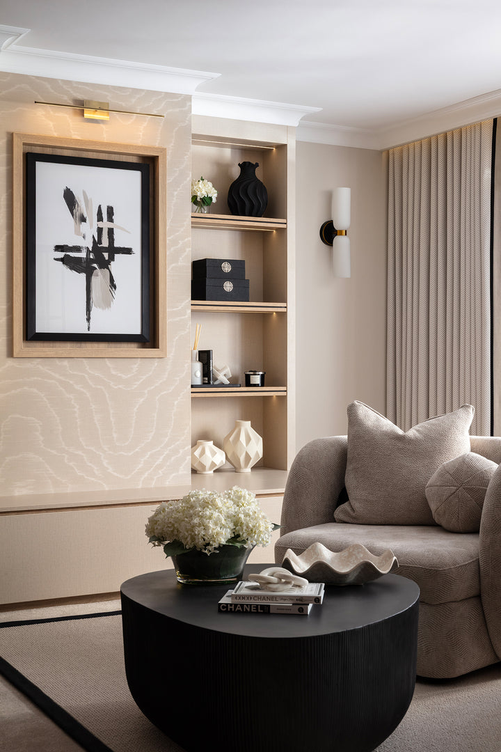 A refined sitting room corner with a dark ribbed drum coffee table, a taupe bouclé armchair, built-in shelving styled with black and cream objects, and a bold black and white abstract artwork - designed by Lisa Hebsy Design & Build Studio