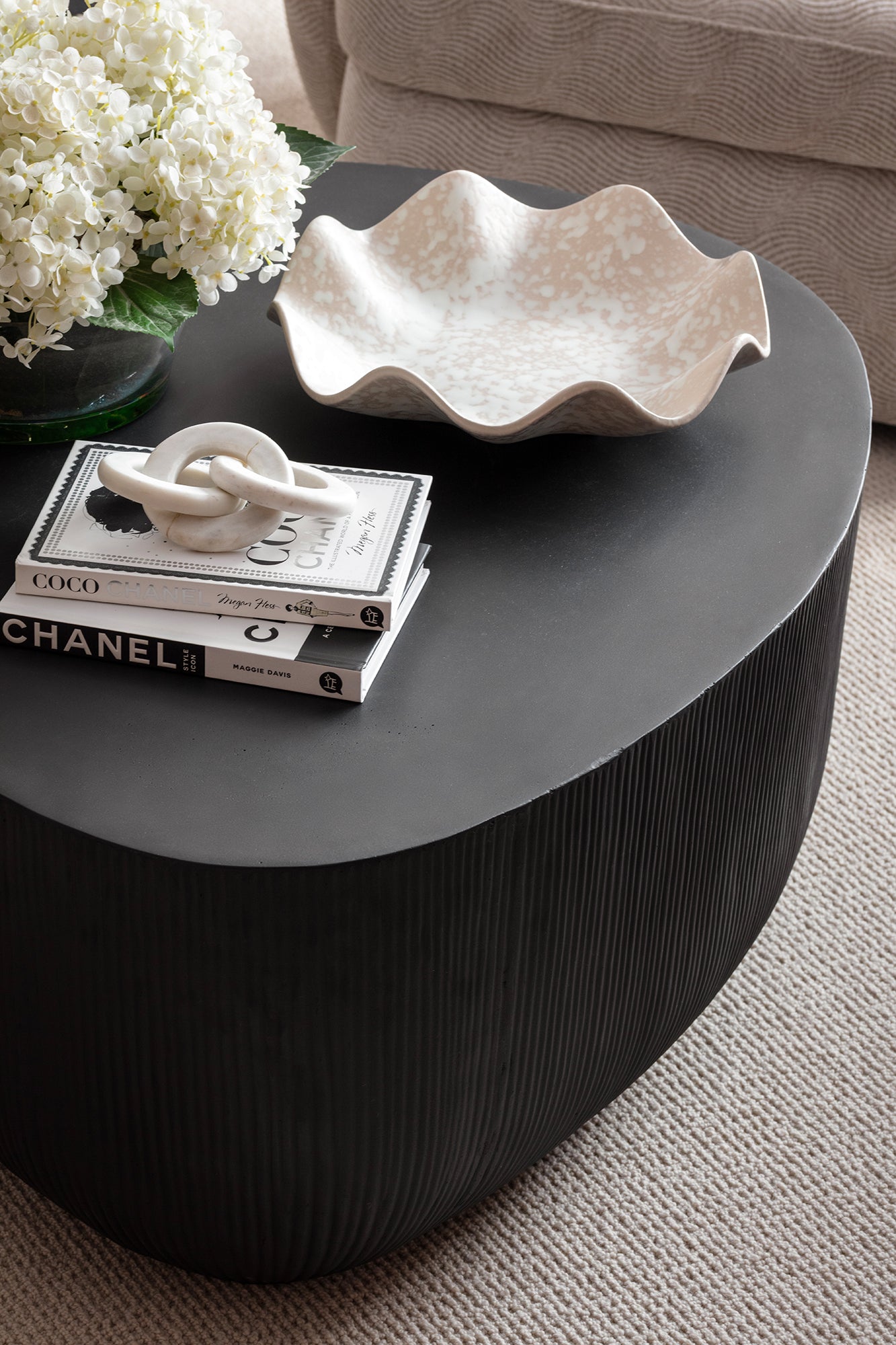 A close-up of a dark ribbed round coffee table styled with fashion books, a white ceramic knot sculpture, a wavy cream dish and a bunch of white hydrangeas - styled by Lisa Hensby Desibgn & Build Studio.