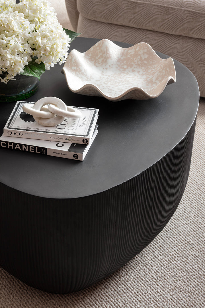 A close-up of a dark ribbed round coffee table styled with fashion books, a white ceramic knot sculpture, a wavy cream dish and a bunch of white hydrangeas - styled by Lisa Hensby Desibgn & Build Studio.