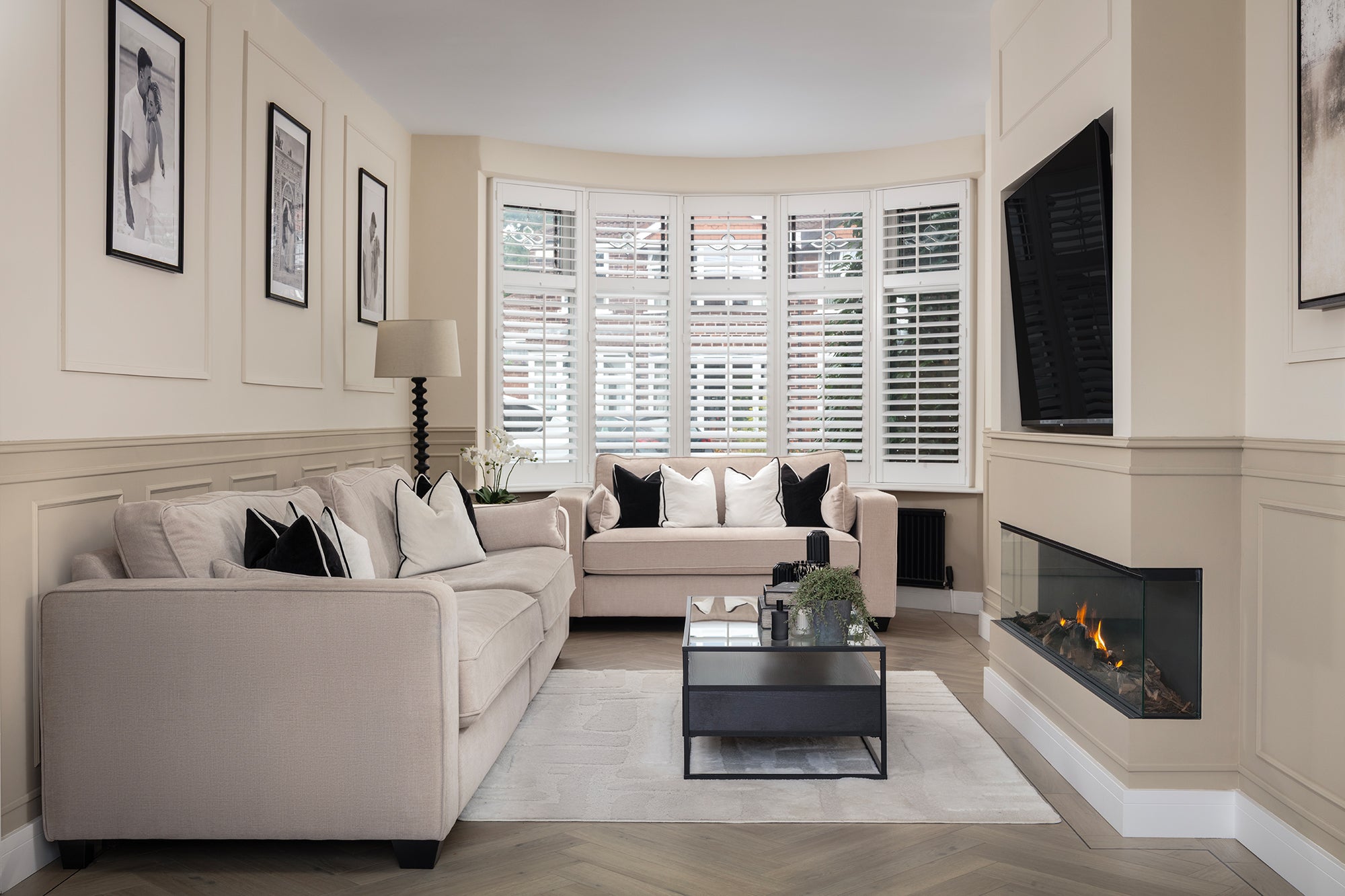 A classic cream living room with a bay window dressed in white plantation shutters, two matching sofas with black and white cushions, a glass coffee table and a wall-mounted gas fireplace — designed by Lisa Hensby Design & Build Studio.