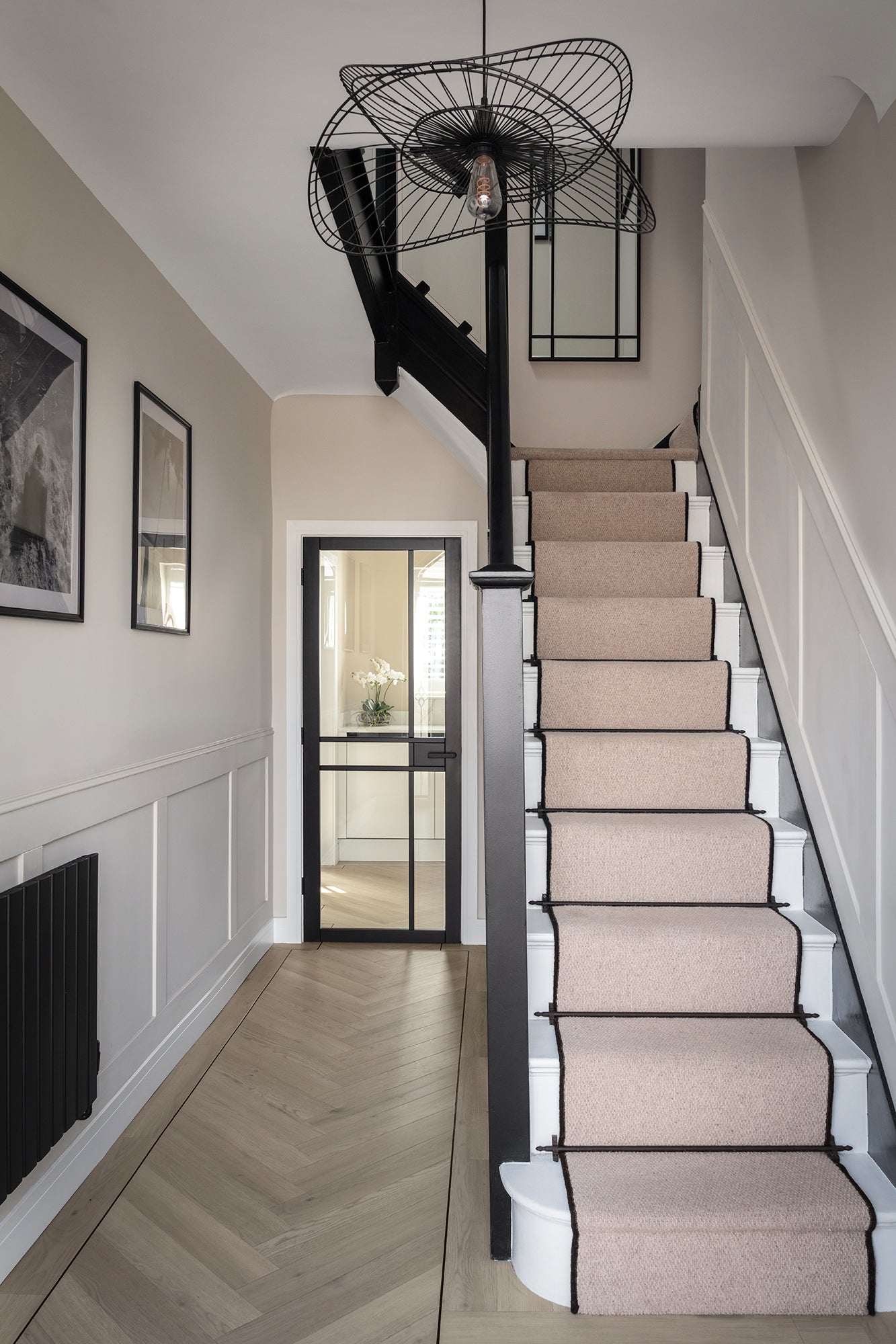 A classic hallway with a light oak herringbone floor, a staircase with a blush pink carpet runner edged in black, black painted banister, white panelled walls and a sculptural black wire pendant light - designed by Lisa Hensby Design & Build Studio.