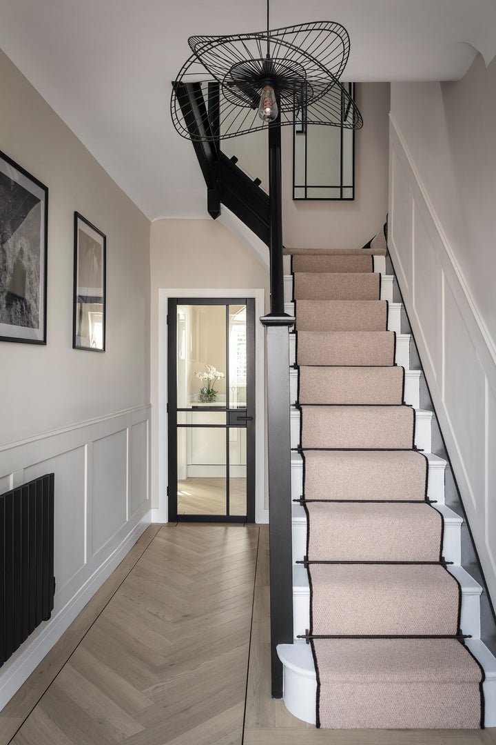 A classic hallway with a light oak herringbone floor, a staircase with a blush pink carpet runner edged in black, black painted banister, white panelled walls and a sculptural black wire pendant light - designed by Lisa Hensby Design & Build Studio.
