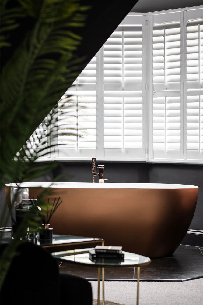 A dramatic dark bedroom featuring a freestanding copper-toned bath with a rose gold floor-mounted tap, white plantation shutters, and lush palm leaves in the foreground —  designed by Lisa Hensby Design & Build Studio.