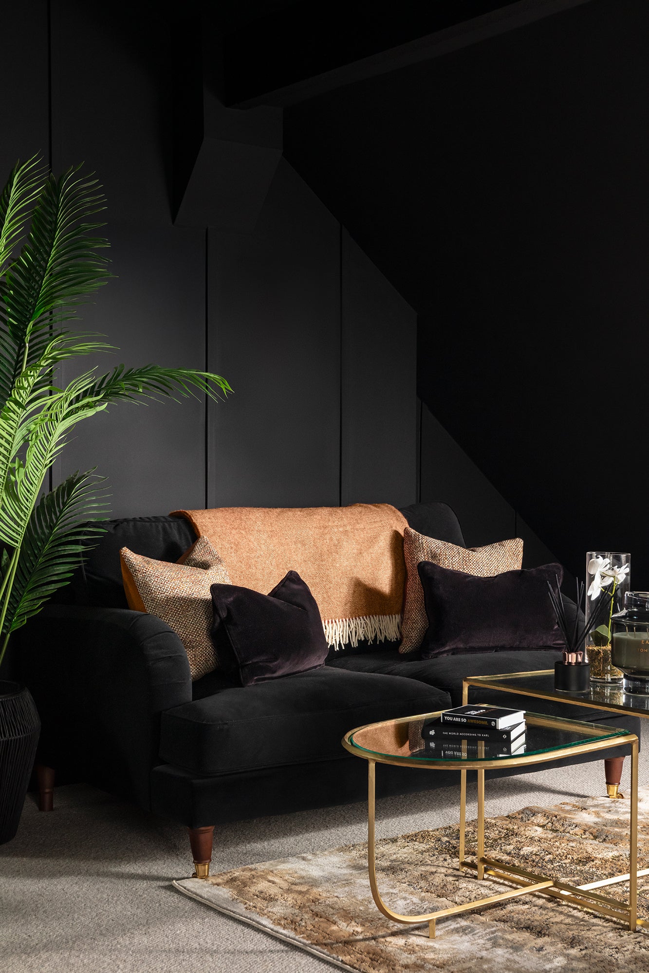 A dramatic dark living room featuring a black velvet sofa styled with copper and dark cushions, a gold-framed glass coffee table, and a tall palm plant against deep charcoal panelled walls - designed and styled by Lisa Hensby Design & Build Studio.