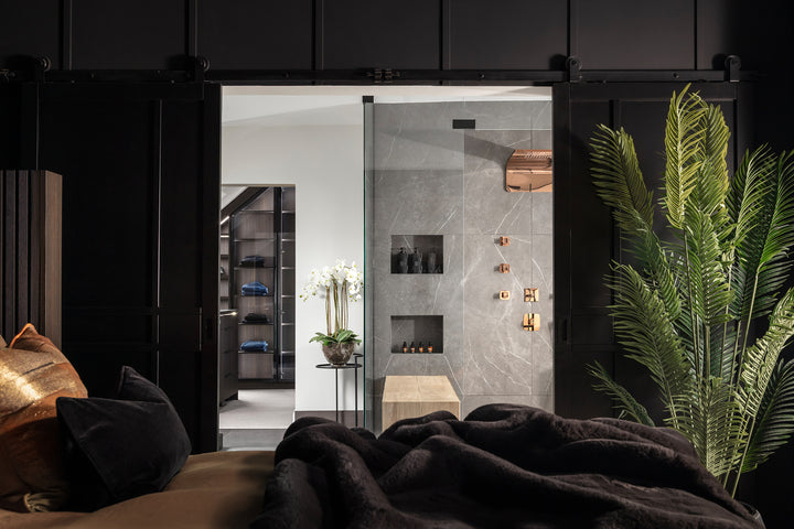 A view from a dark bedroom through an open sliding barn door into a luxury ensuite shower room with grey marble-effect tiles, rose gold fittings, and a walk-in dressing room beyond - designed by Lisa Hensby Design & Build Studio.