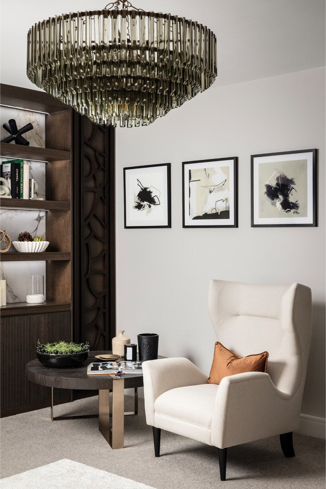 A refined sitting room corner with a cream wingback armchair, a trio of abstract artworks, a large smoked glass chandelier and dark walnut fitted shelving with decorative lattice detail - styled and designed by Lisa Hensby Design & Build Studio.