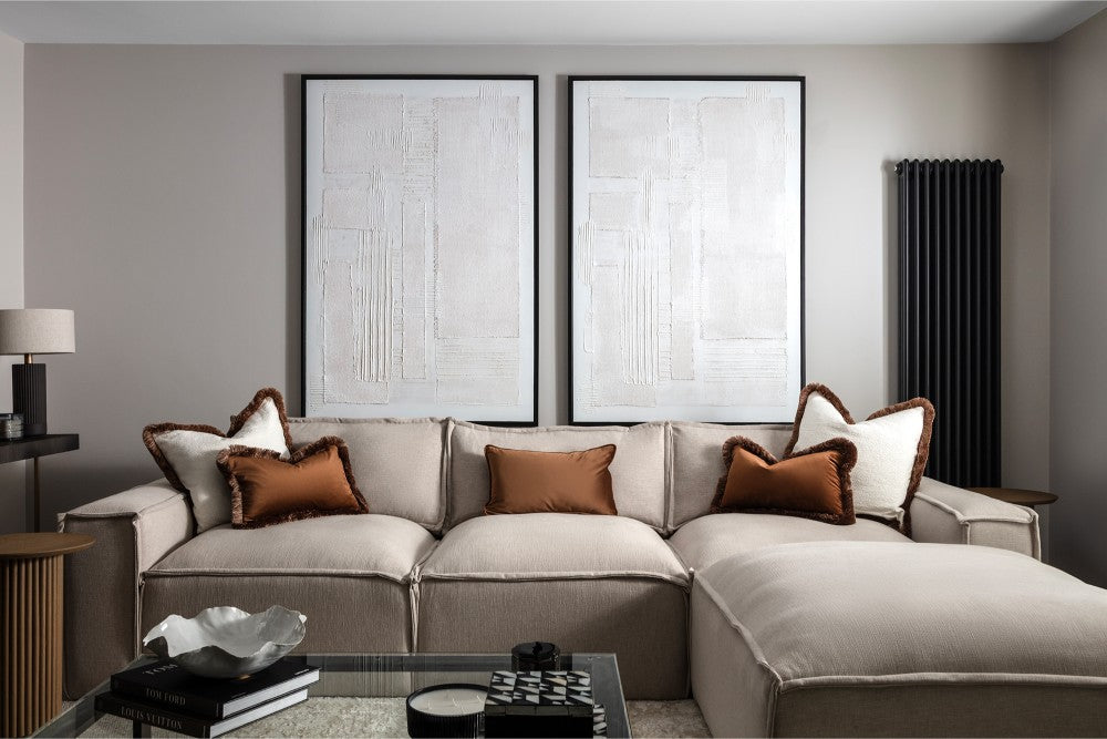 A neutral living room with a large cream corner sofa styled with rust and white fringe-edged cushions, a pair of oversized white textured artworks in black frames, and a glass coffee table — designed by Lisa Hensby Design & Build Studio.