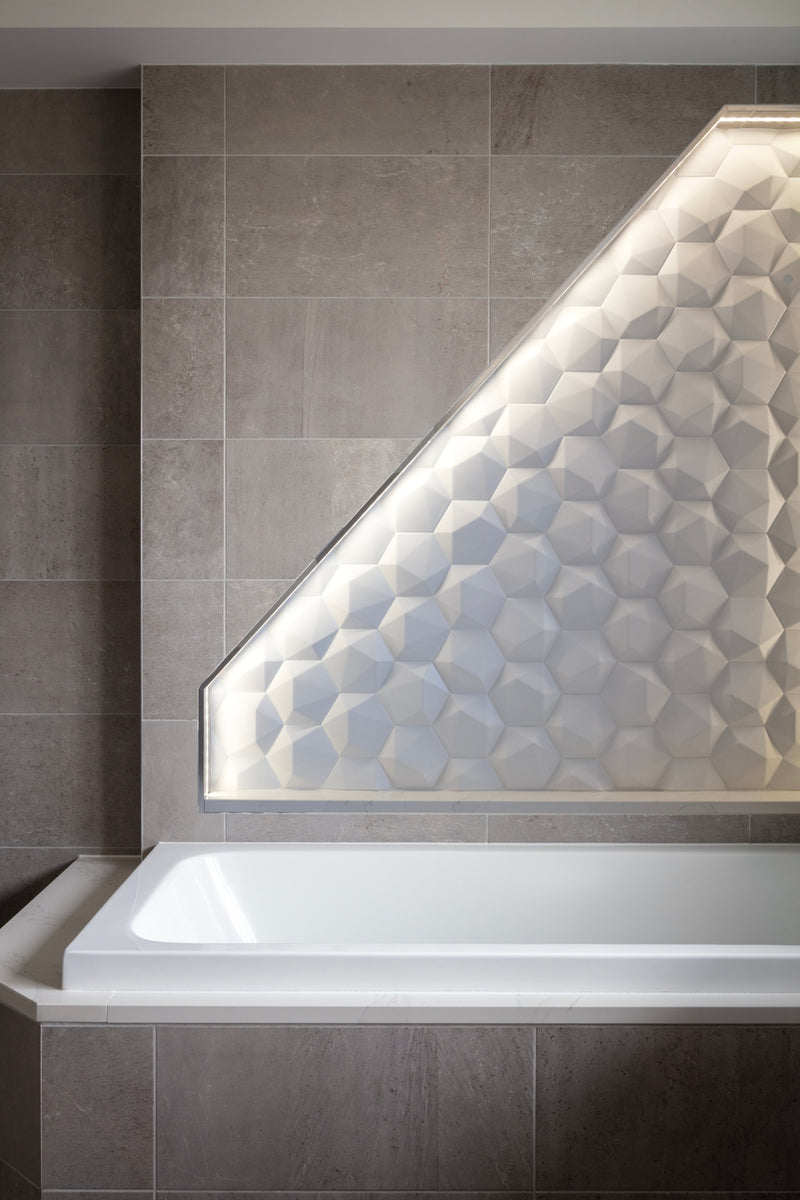 Built-in bath beneath a backlit white 3D geometric tile wall set into warm grey tiles, in Lisa Hensby's own bathroom