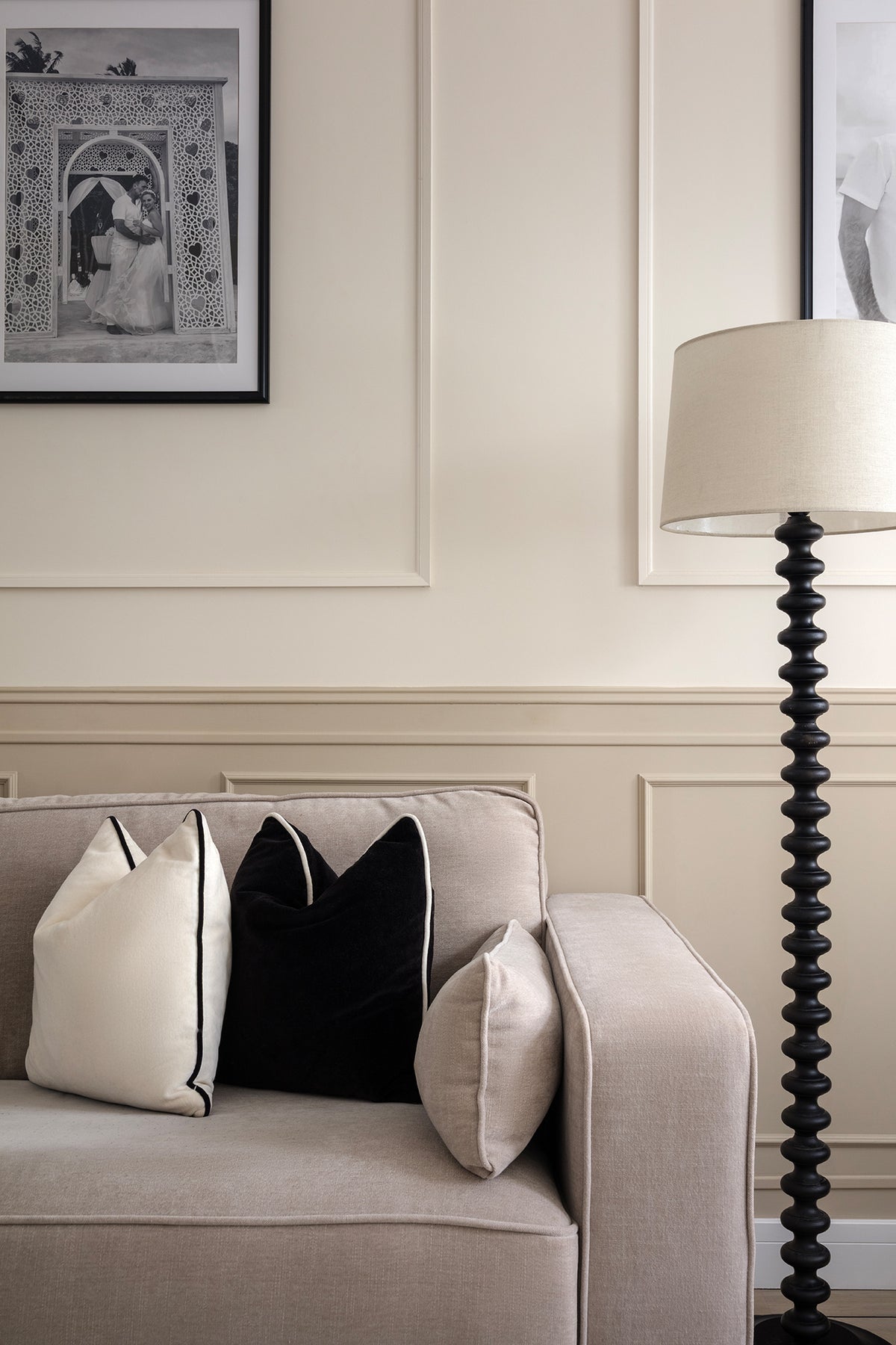 Neutral linen sofa styled with white and black cushions, set against panelled walls with black and white framed photography and a sculptural black turned-wood floor lamp. Designed by Lisa Hensby Design & Build Studio.