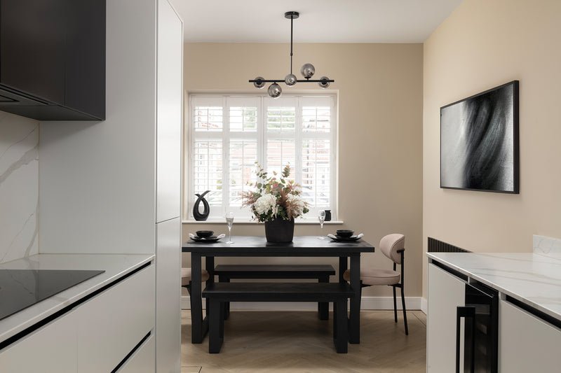 Contemporary monochrome dining nook with a black wood table and benches, cream accent chair, globe pendant light, plantation shutters and herringbone flooring, adjoining a white marble-effect kitchen. Designed by Lisa Hensby Design & Build Studio.