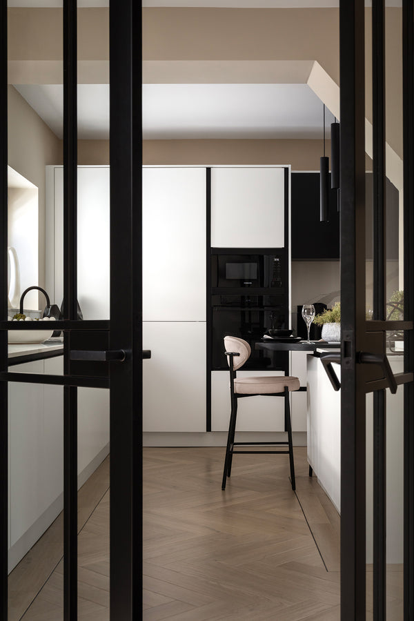 Black Crittall-style glazed doors framing a view into a monochrome kitchen with white handleless cabinetry, integrated black appliances, a cream bar stool and herringbone flooring. Designed by Lisa Hensby Design & Build Studio.