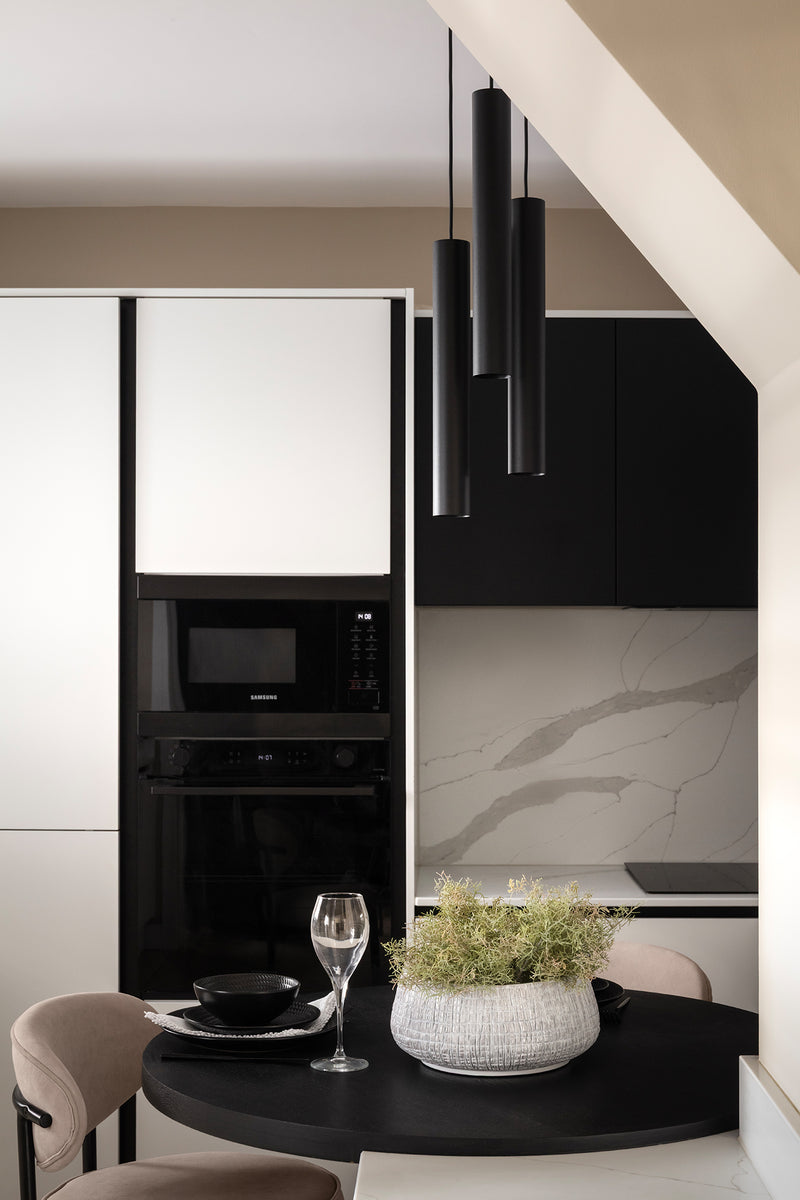 Round black dining table with cream chairs and styled greenery, set against white and black handleless cabinetry, integrated black appliances, a marble-effect splashback and slim black pendant lights. Designed by Lisa Hensby Design & Build Studio.