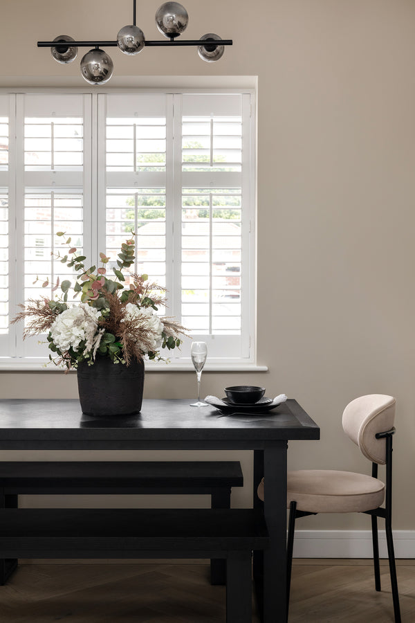 Black wood dining table and bench with a cream accent chair, styled with a floral centrepiece and black place setting, beneath plantation shutters and a black globe pendant light. Designed by Lisa Hensby Design & Build Studio.