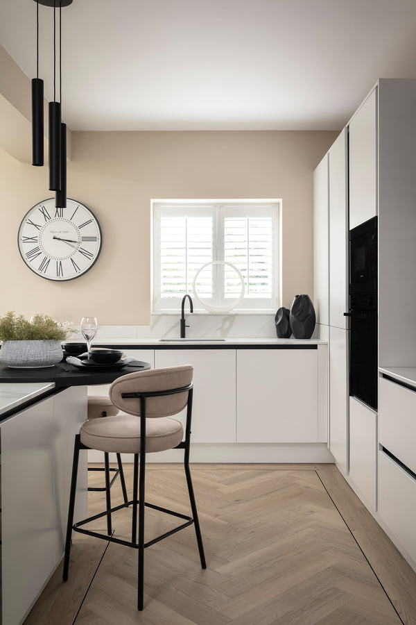 White handleless kitchen with integrated black appliances, a cream bar stool at a black breakfast bar, plantation-shuttered window, a large round wall clock and herringbone flooring. Designed by Lisa Hensby Design & Build Studio.