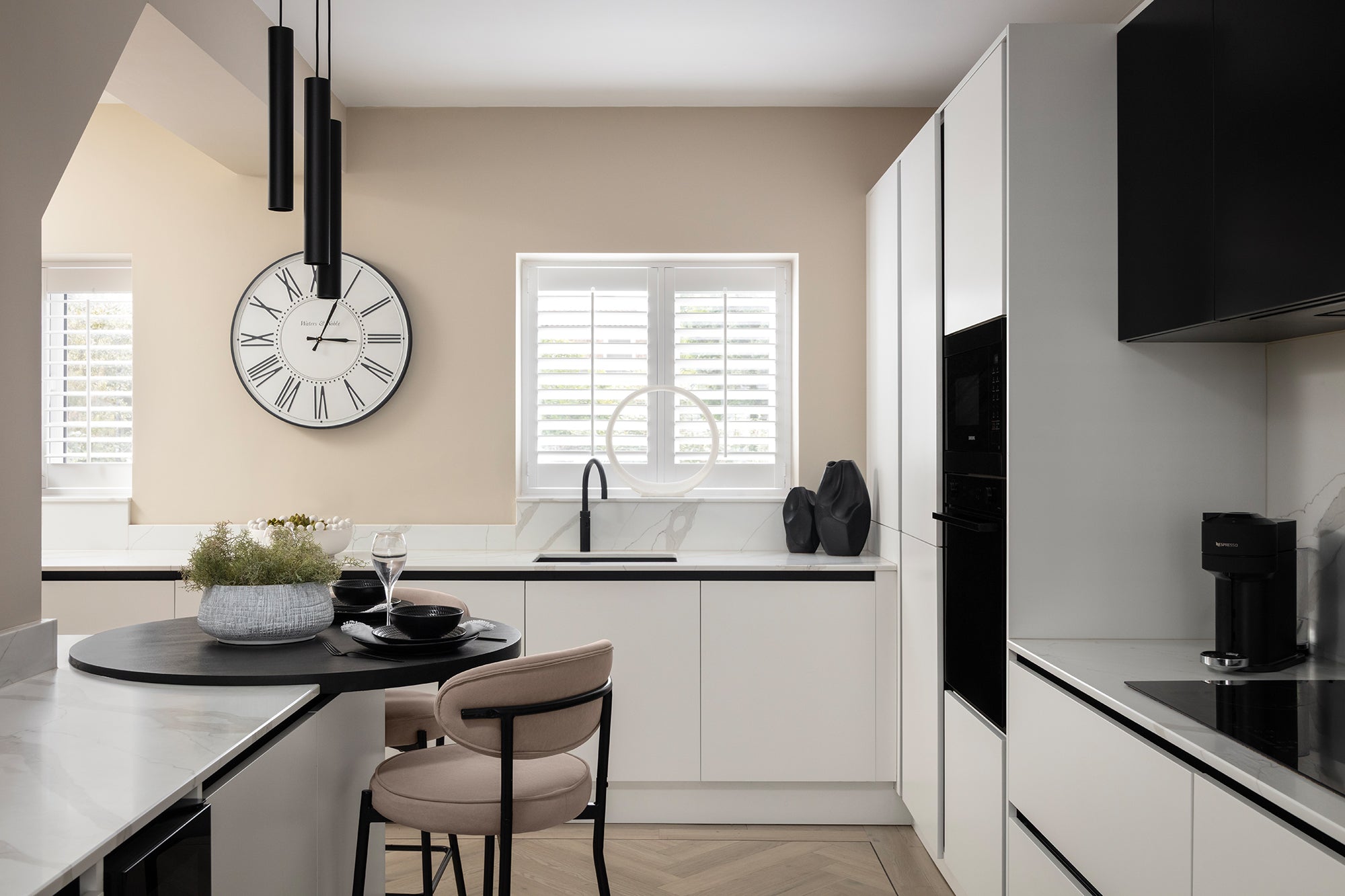 Monochrome kitchen with white and black cabinetry, integrated black appliances, a round breakfast bar with cream chairs, marble-effect splashback, a large wall clock and herringbone flooring. Designed by Lisa Hensby Design & Build Studio.