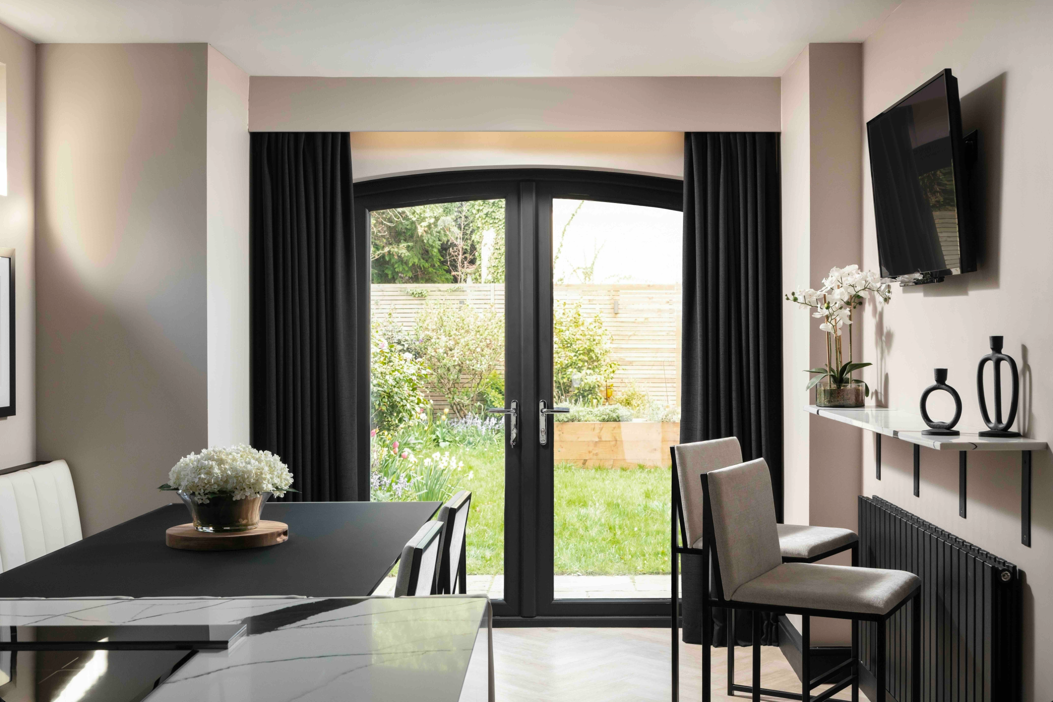 A modern dining room with black French doors opening onto a sunny garden, flanked by black curtains, with a marble-effect dining table and bar-height seating - designed by Lisa Hensby Design & Build Studio.