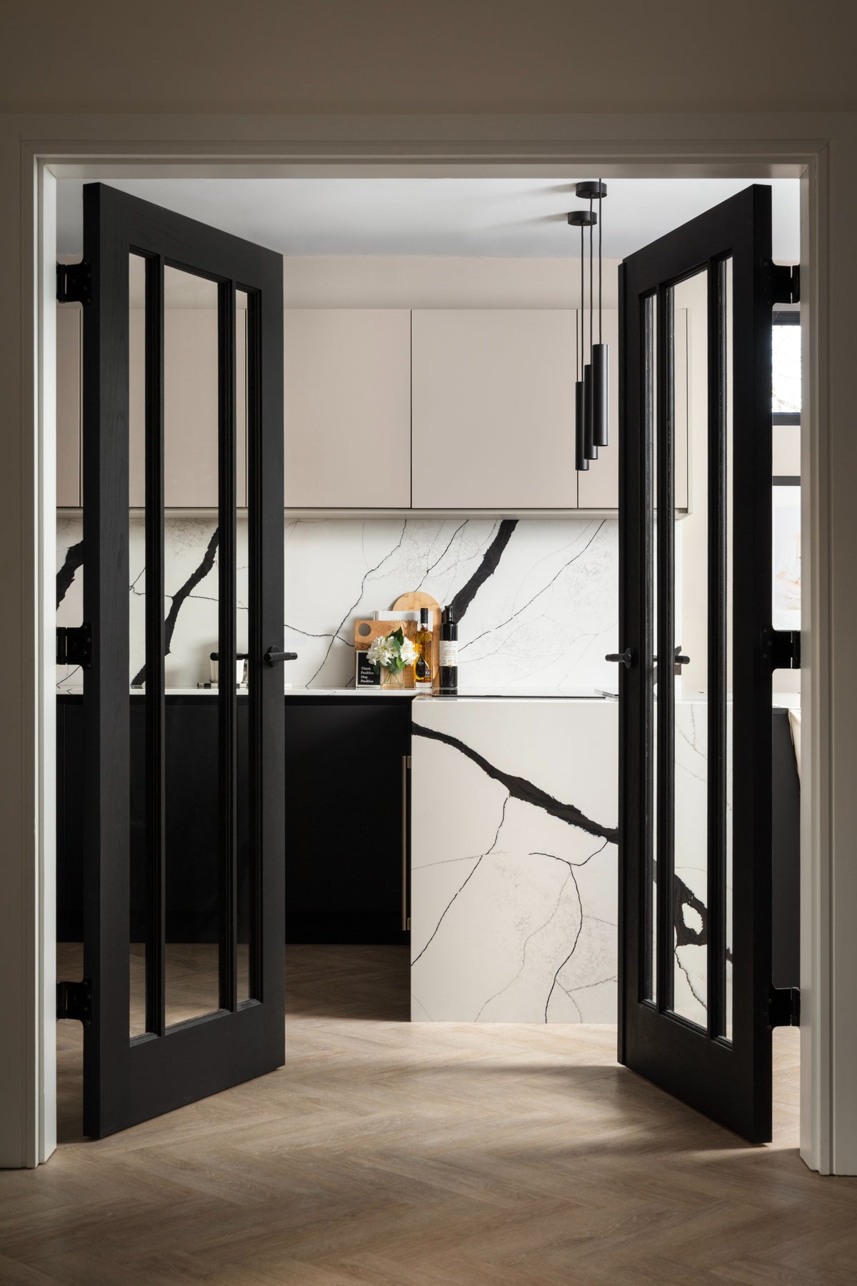 Black glass-panelled doors open onto a kitchen with a black quartz waterfall island, cashmere wall units and herringbone flooring. Designed by Lisa Hensby Design & Build Studio