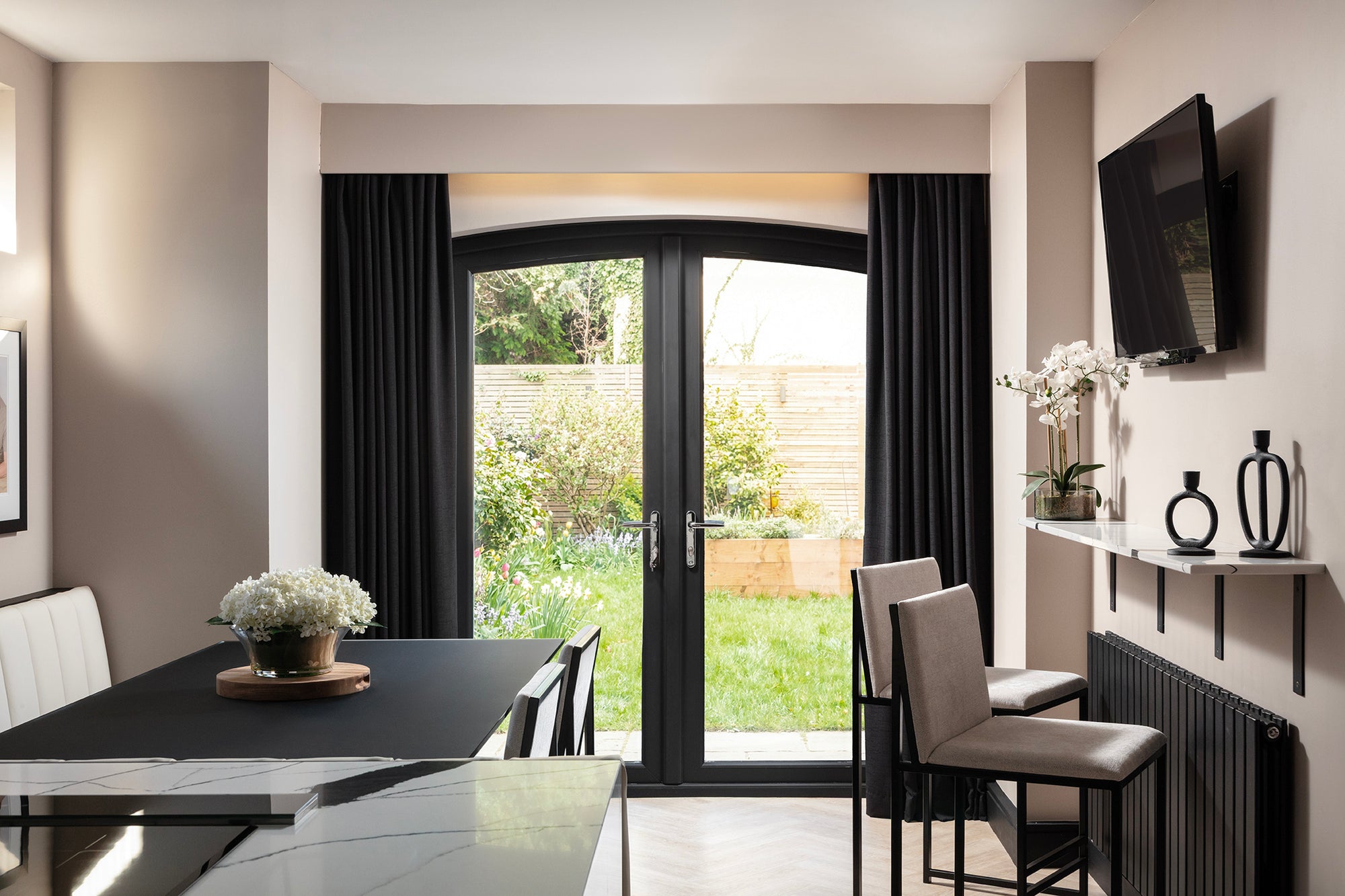 Kitchen-diner opening onto the garden through arched French doors with charcoal curtains, alongside the dining table, a floating stone shelf and the quartz island. Designed by Lisa Hensby Design & Build Studio.