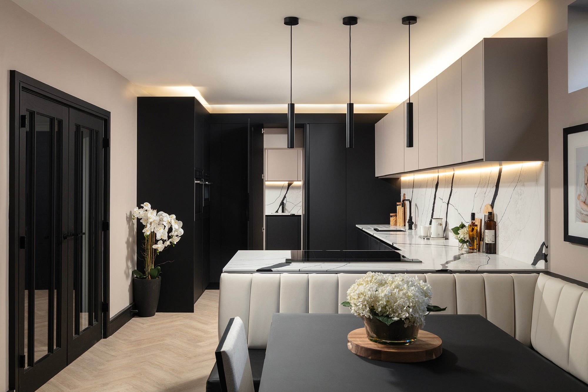 Long view of the kitchen-diner from glass-panelled doors through jet black units, cashmere wall units and quartz island to the cream banquette booth and dining table. Designed by Lisa Hensby Design & Build Studio.