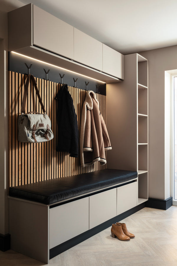 A bespoke hallway boot room with greige fitted cabinetry, a vertical oak slat back panel with black coat hooks, a black leather bench seat with storage below, and a herringbone oak floor - designed by Lisa Hensby Design & Build Studio.