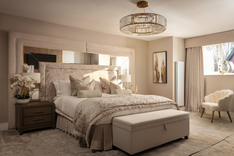 A luxurious neutral bedroom with a tall upholstered headboard, large mirror, layered champagne bedding, a gold crystal chandelier, and a cream bouclé armchair in the corner. Designed by Lisa Hensby Design & Build Studio.