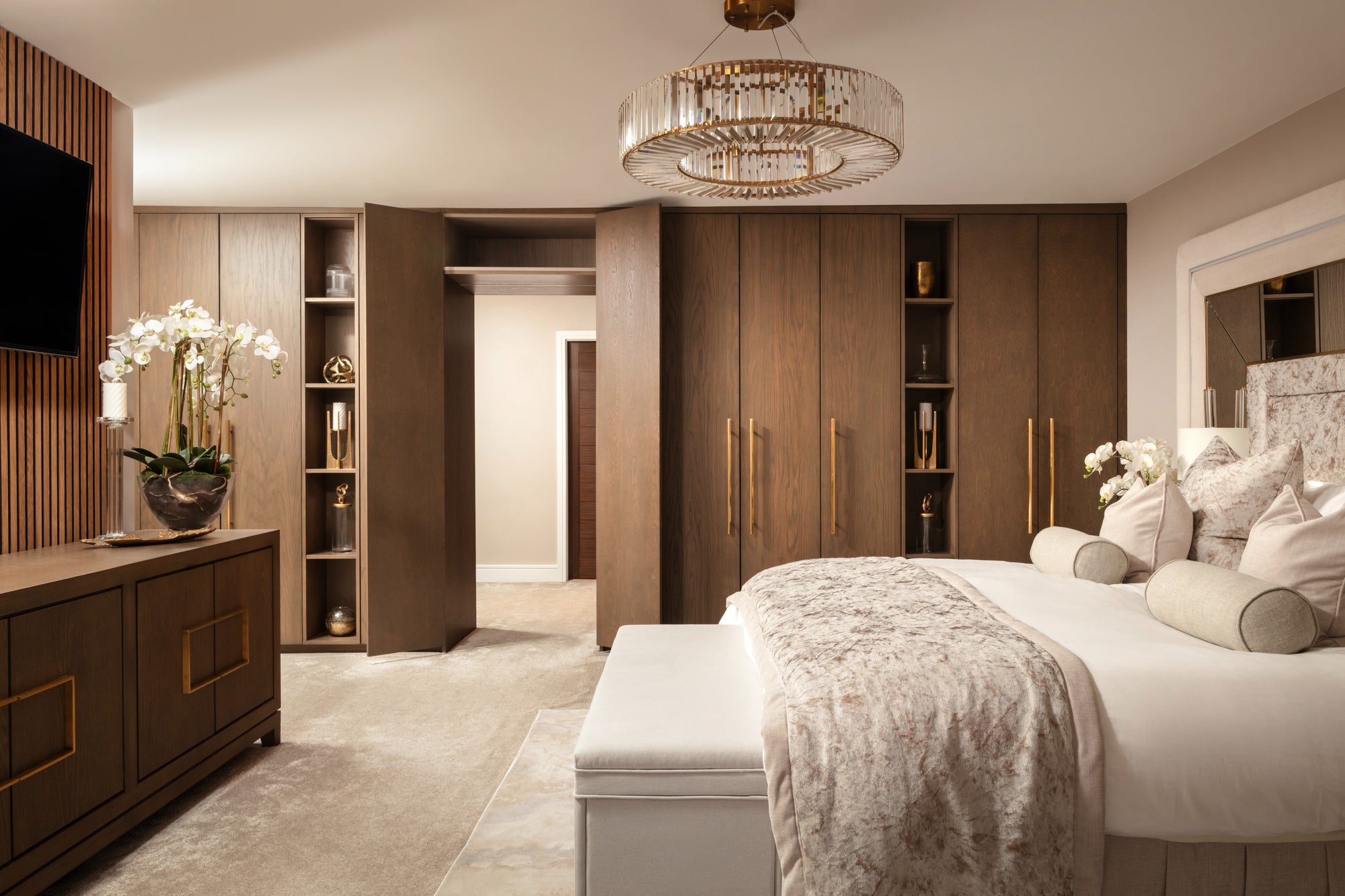 Master bedroom with dark oak fitted wardrobes and gold handles, statement crystal chandelier, dressed bed in champagne crushed velvet with matching upholstered bench, and a wood-panelled wall with wall-mounted TV. Designed by Lisa Hensby Design & Build Studio. 