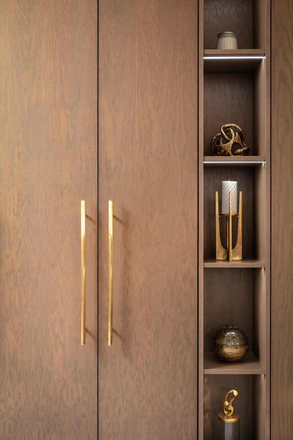 Close-up of dark oak wardrobe doors with slim brass handles, beside an open shelving column styled with brass ornaments, a stone candle holder and a glass vase, lit with integrated LED strip lighting. Designed by Lisa Hensby Design & Build Studio. 