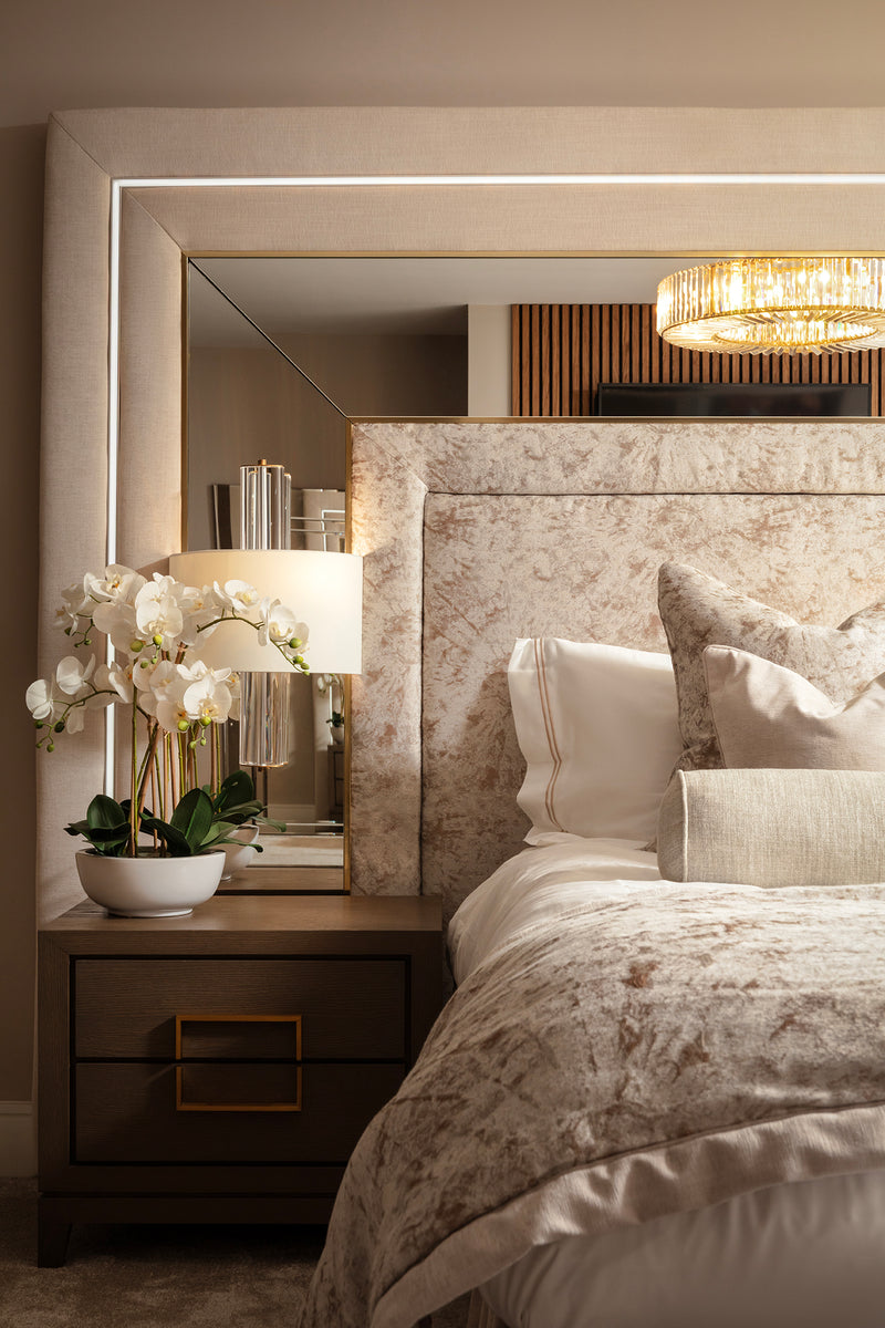 Bedside detail with backlit mirror, dark wood nightstand with brass-edged drawer, table lamp and white orchid, next to a crushed velvet upholstered headboard and matching bedding. Designed by Lisa Hensby Design & Build Studio.