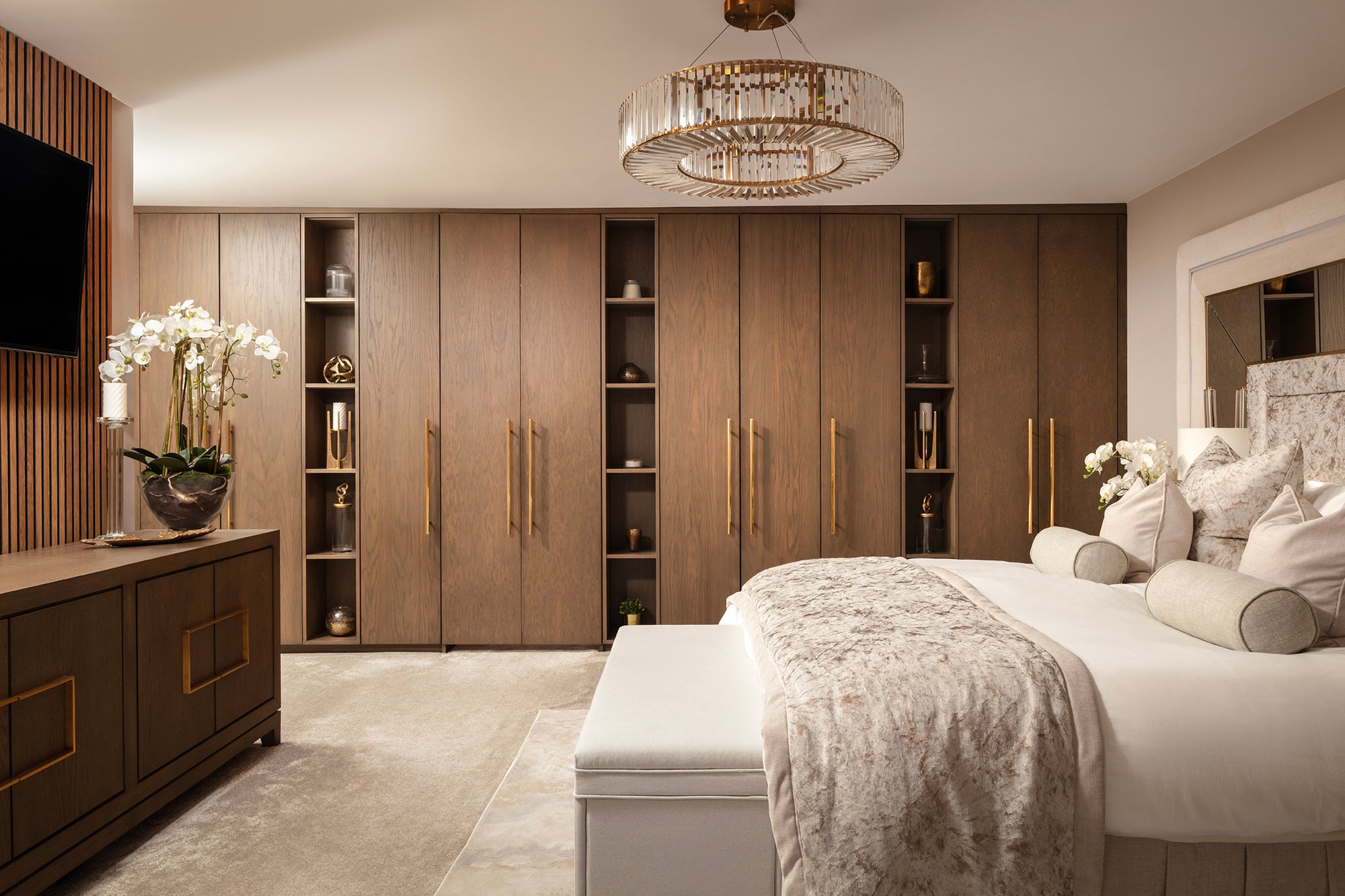 Full-width bedroom shot showing a run of dark oak fitted wardrobes with open shelving and brass handles, a crystal drum chandelier, and a bed dressed in champagne crushed velvet with a matching upholstered bench. Designed by Lisa Hensby Design & Build Studio.