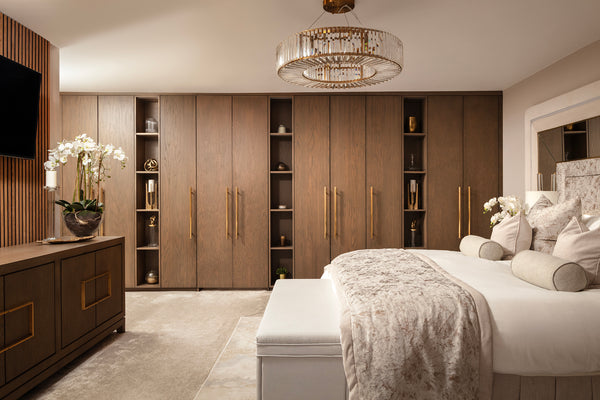 Full-width bedroom shot showing a run of dark oak fitted wardrobes with open shelving and brass handles, a crystal drum chandelier, and a bed dressed in champagne crushed velvet with a matching upholstered bench. Designed by Lisa Hensby Design & Build Studio.