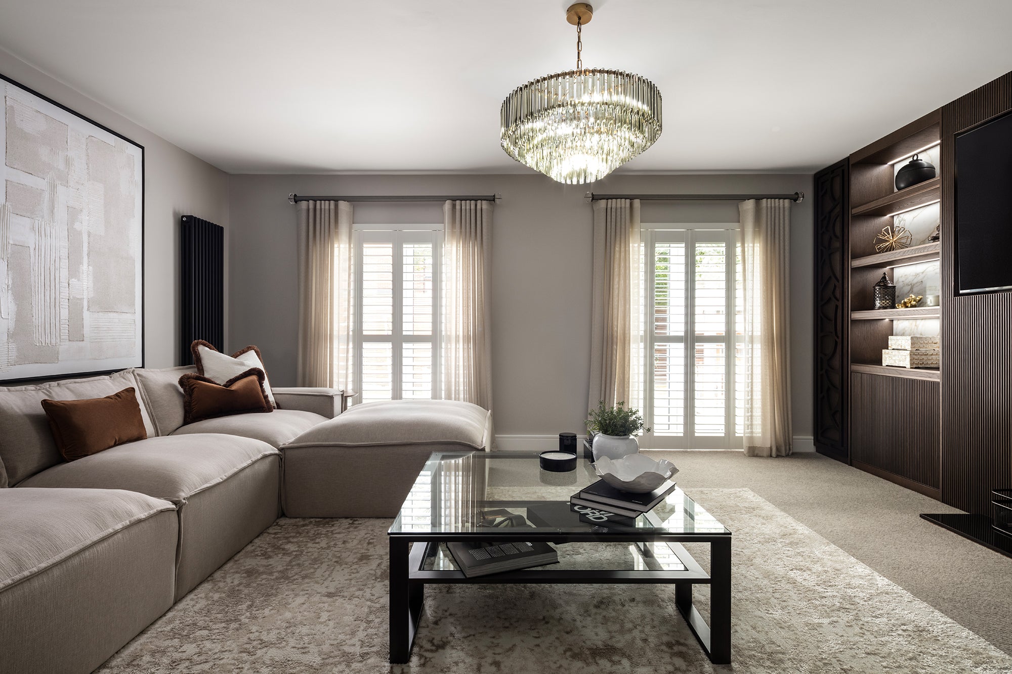 
Neutral, elegant living room with a beige L-shaped sectional, black glass coffee table, crystal chandelier, plantation-shutter windows and a dark wood built-in shelving unit. Designed by Lisa Hensby of Lisa Hensby Design & Build Studio.