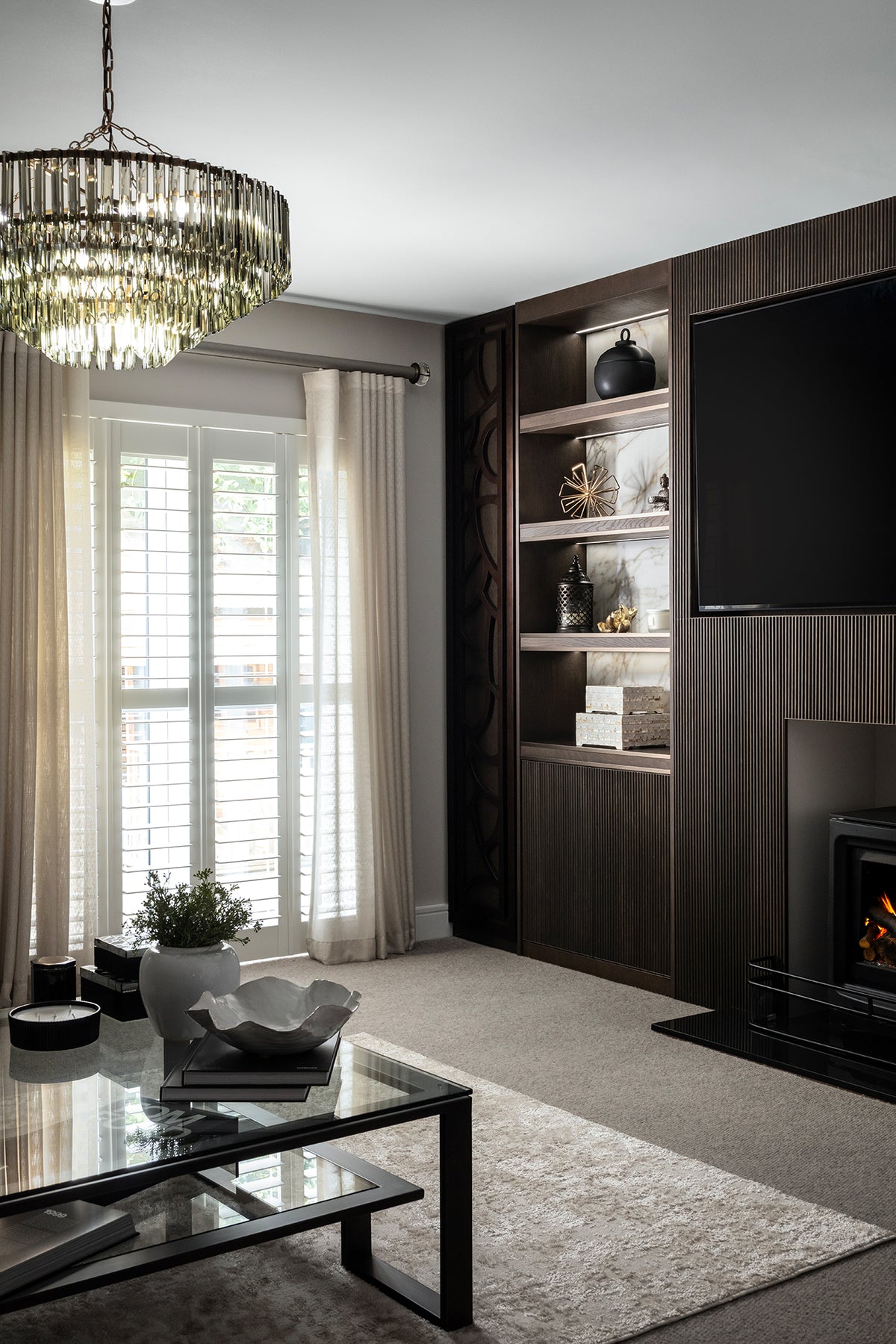 Elegant living room with a dark fluted wood media wall featuring a wall-mounted TV, built-in shelving and a fireplace, alongside a glass-topped coffee table, neutral rug, plantation shutters and sheer curtains, lit by a tiered smoked-glass chandelier. Designed Lisa Hensby Design & Build Studio.