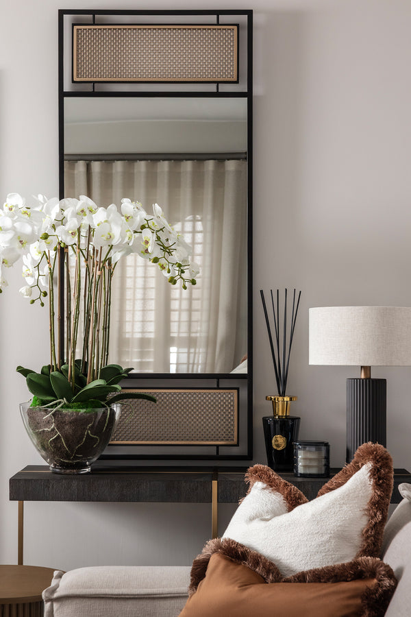 A styled console-table vignette in a softly lit, neutral interior: a tall black-framed mirror with woven cane panels reflects sheer curtains, above a dark wood console with brass legs. To the left, white orchids arch out of a marbled black bowl; to the right sit a black reed diffuser, a candle, and a fluted dark table lamp with a cream shade. A cream cushion with a brown fur trim rests on a sofa in the foreground. Designed by Lisa Hensby of Lisa Hensby Design & Build Studio.