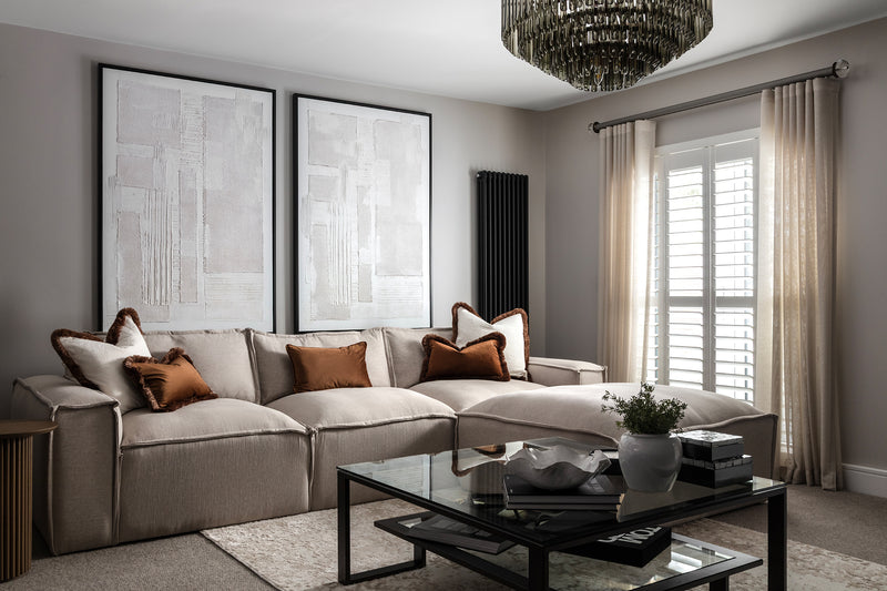 Elegant neutral living room with a beige sectional sofa, rust-brown cushions, a black glass coffee table, twin abstract canvases and a plantation-shutter window beneath a smoked-glass chandelier. Designed by Lisa Hens by of Lisa Hensby Design & Build Studio.
