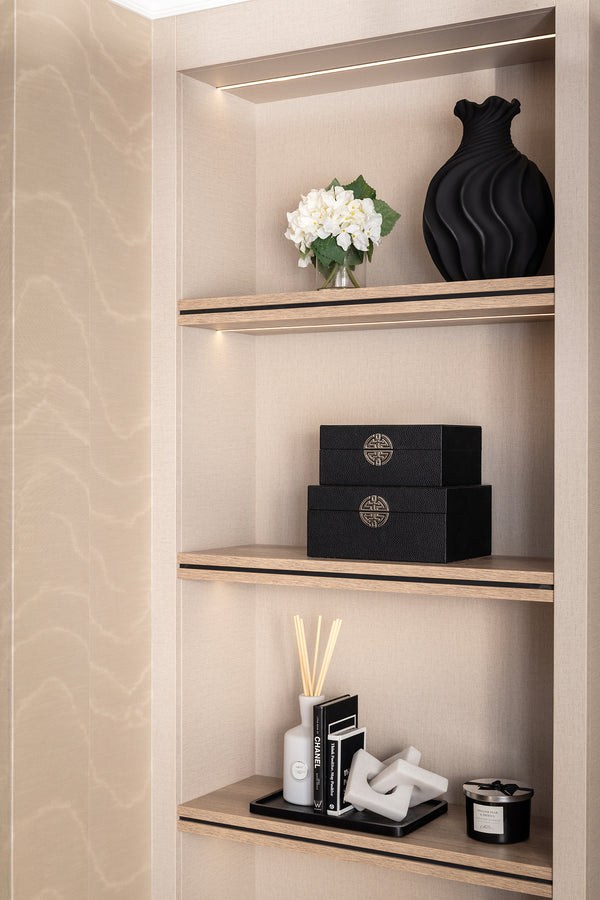 Built-in shelving niche with integrated LED lighting and walnut-effect shelves, styled with a black sculptural vase, white flowers, decorative boxes, candles and a diffuser. Designed by Lisa Hensby of Lisa Hensby Design & Build Studio